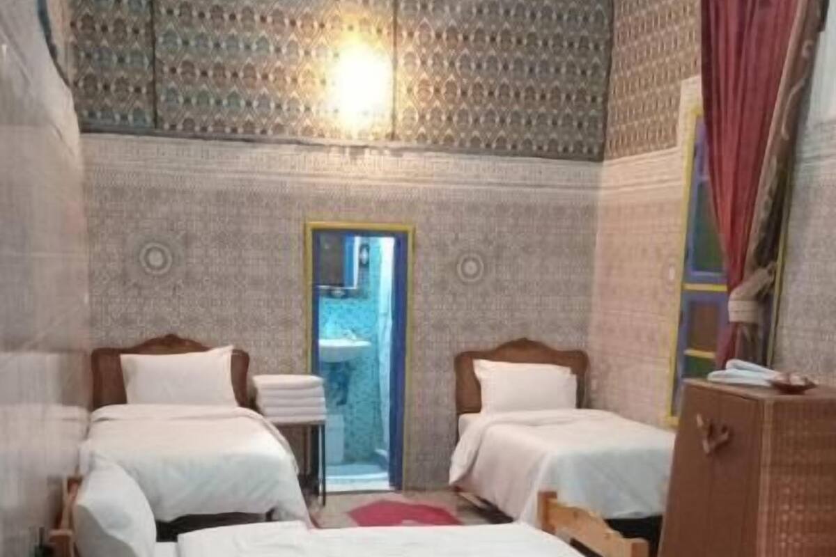 Una habitación con cuatro camas dispuestas en fila, un armario de madera y un papel tapiz estampado, con una puerta que da a un baño.