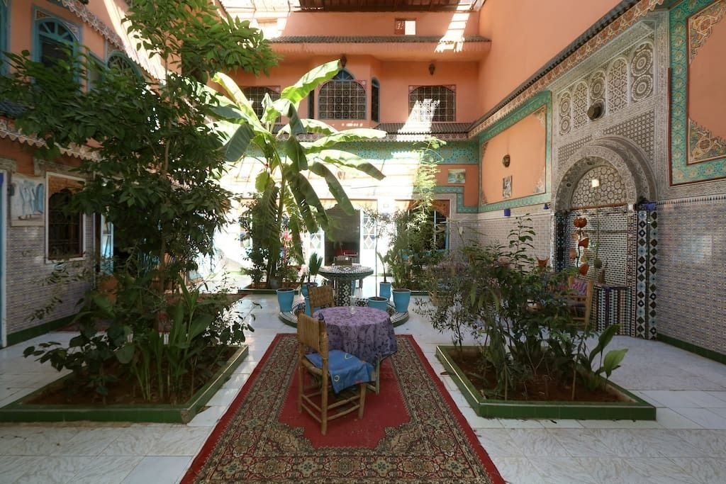 A courtyard with a central round table, surrounded by potted plants and a red carpet.