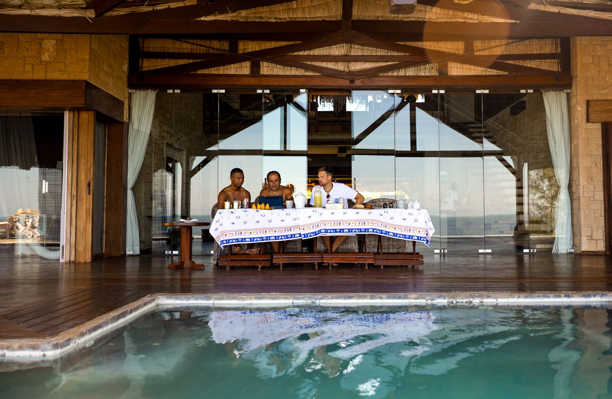 A poolside dining area with a table set for three, surrounded by a glass enclosure, and a clear view of the pool.