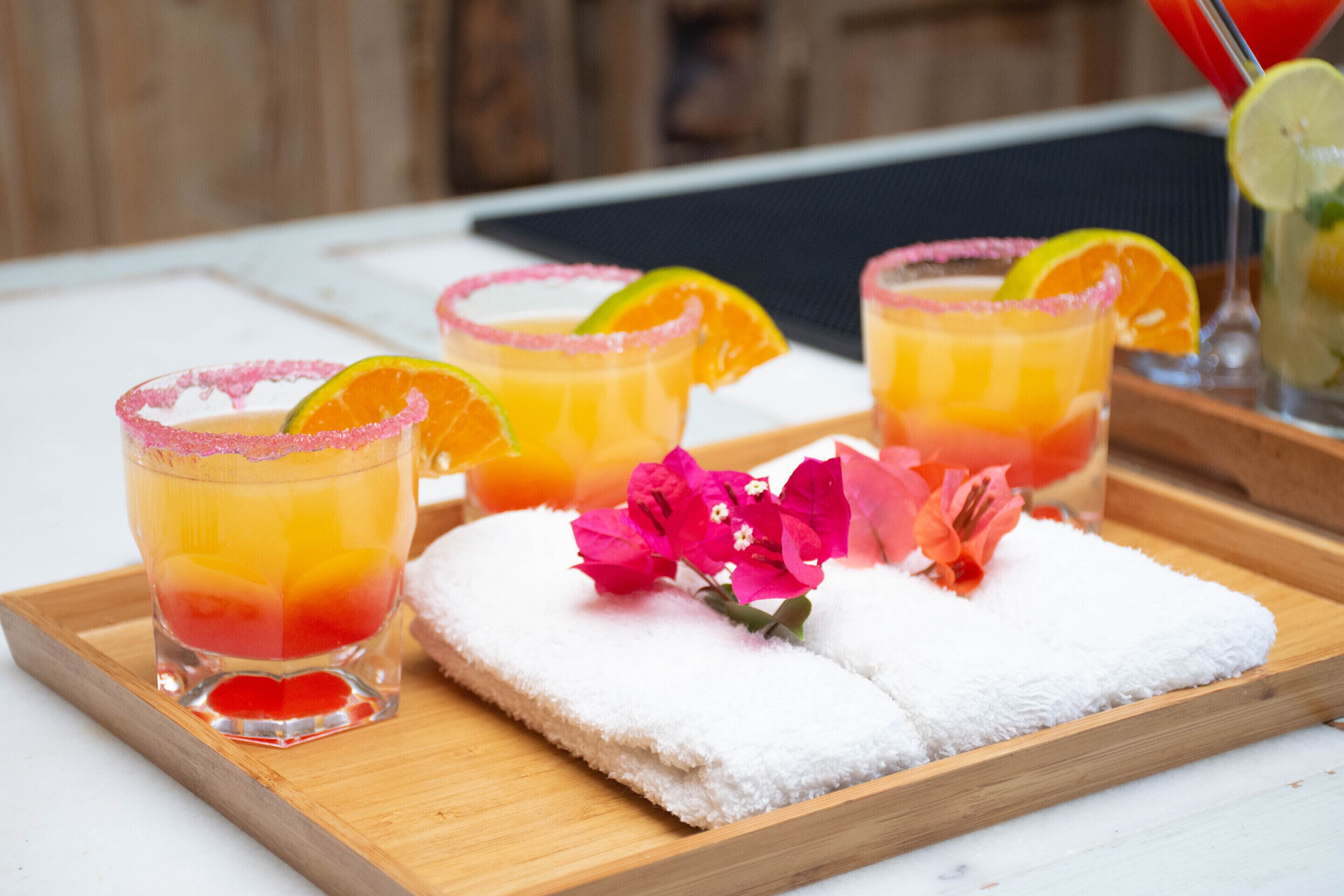 A tray with three glasses of orange juice, each garnished with a slice of lime and a pink rim, and a white towel with pink flowers.