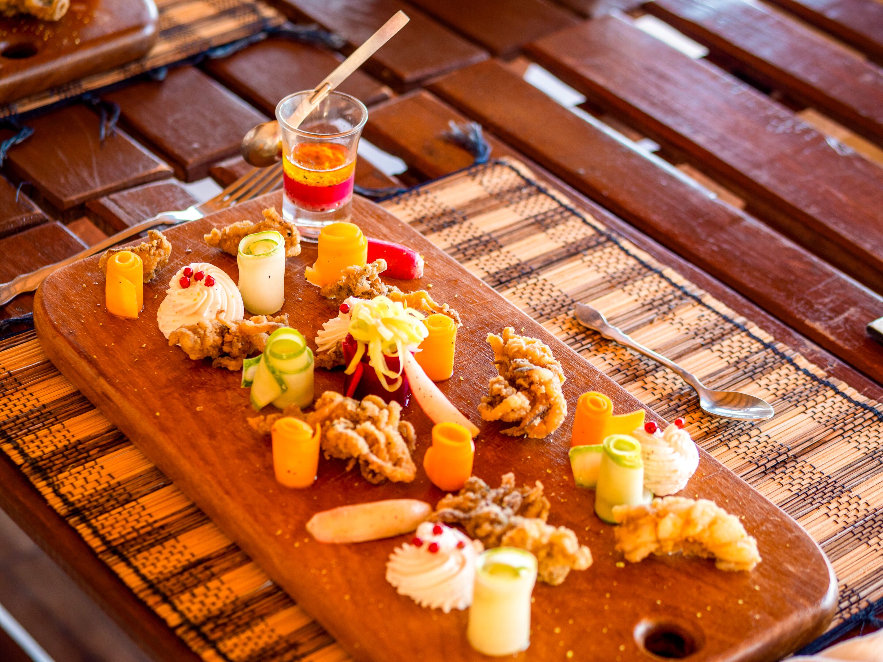 A wooden serving board with various food items and a drink on a woven placemat.
