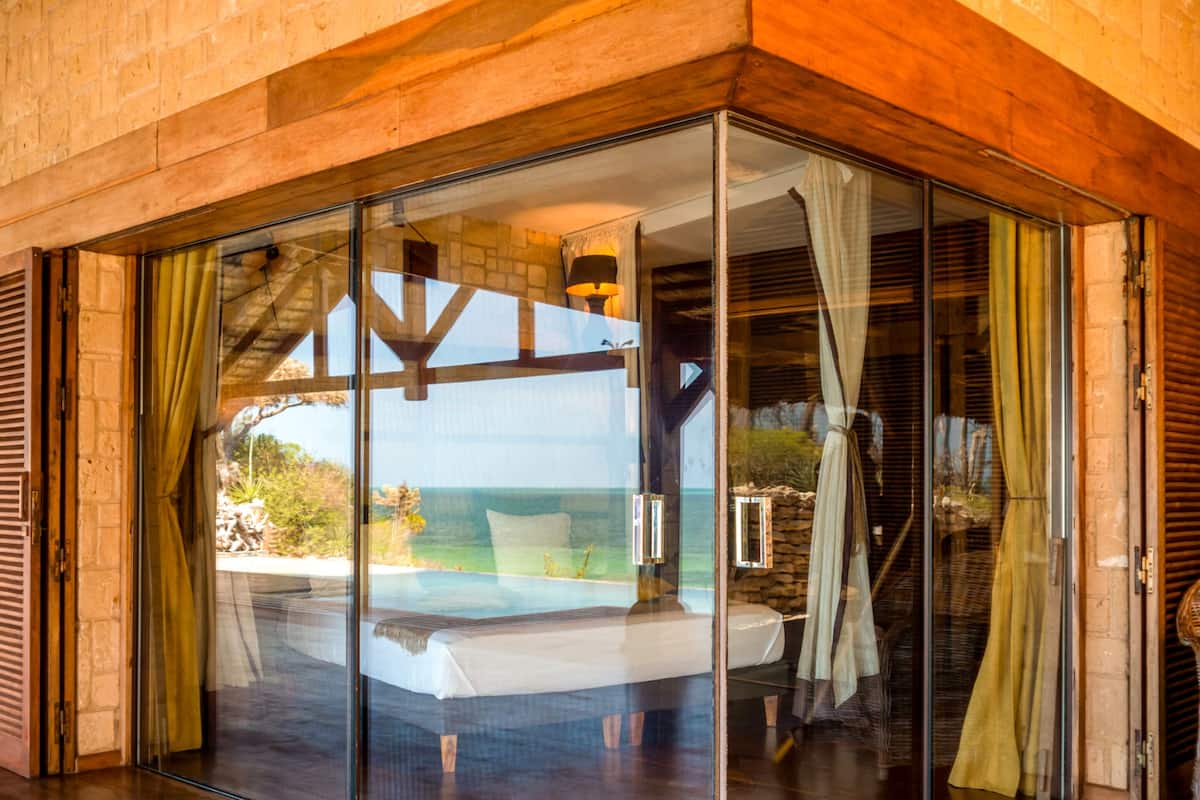 Quarto com paredes de vidro e vista para o oceano, vigas de madeira e parede de pedra.