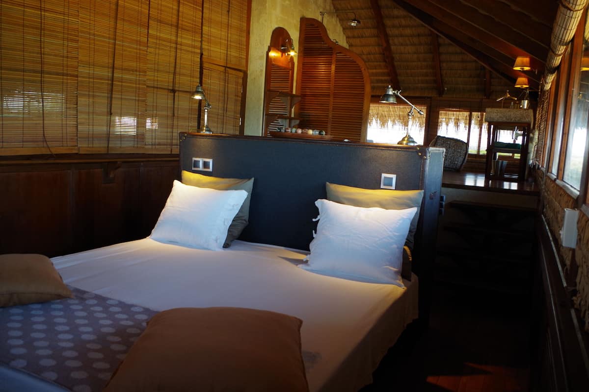 Quarto de hotel com uma cama grande, cabeceira de madeira e uma mesinha com luminária.