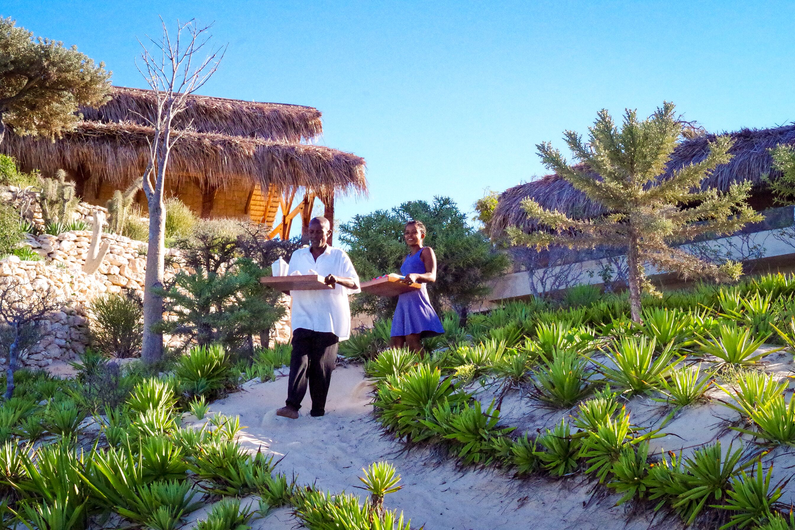 A couple walking on a sandy path surrounded by green plants and thatched-roof structures.