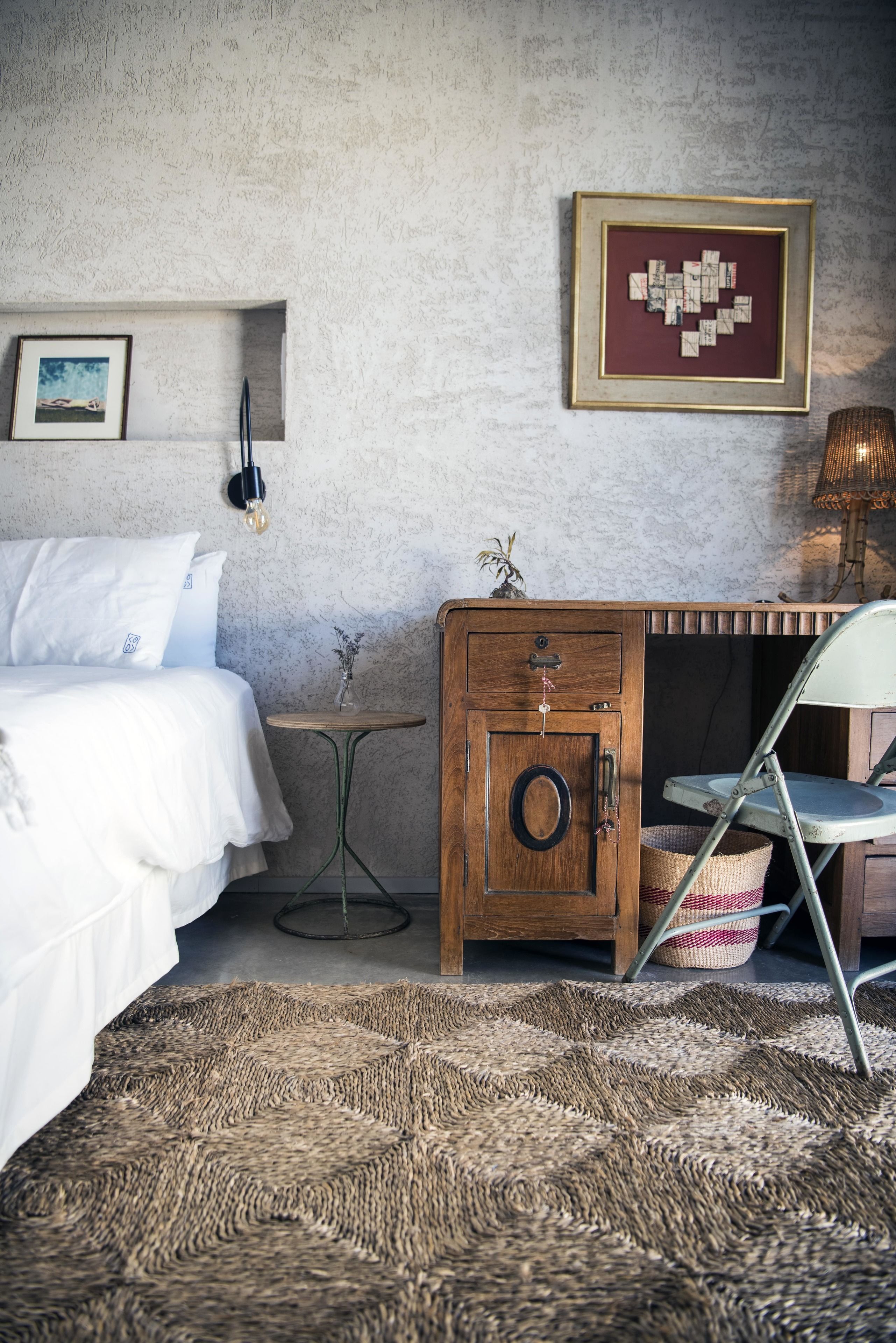 A bedroom with a bed, a wooden nightstand, a small table, a chair, a wall-mounted lamp, and a framed picture.