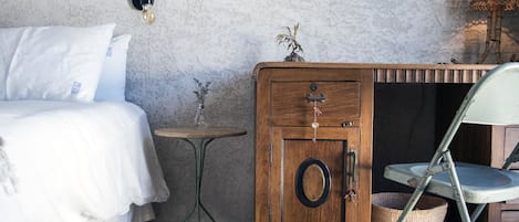 A bedroom with a bed, a wooden nightstand, a small table, a chair, a wall-mounted lamp, and a framed picture.