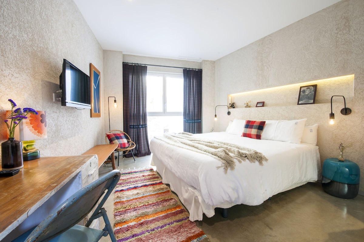 Habitación de hotel moderna con una cama grande, un escritorio con silla, un televisor montado en la pared y una ventana con cortinas.