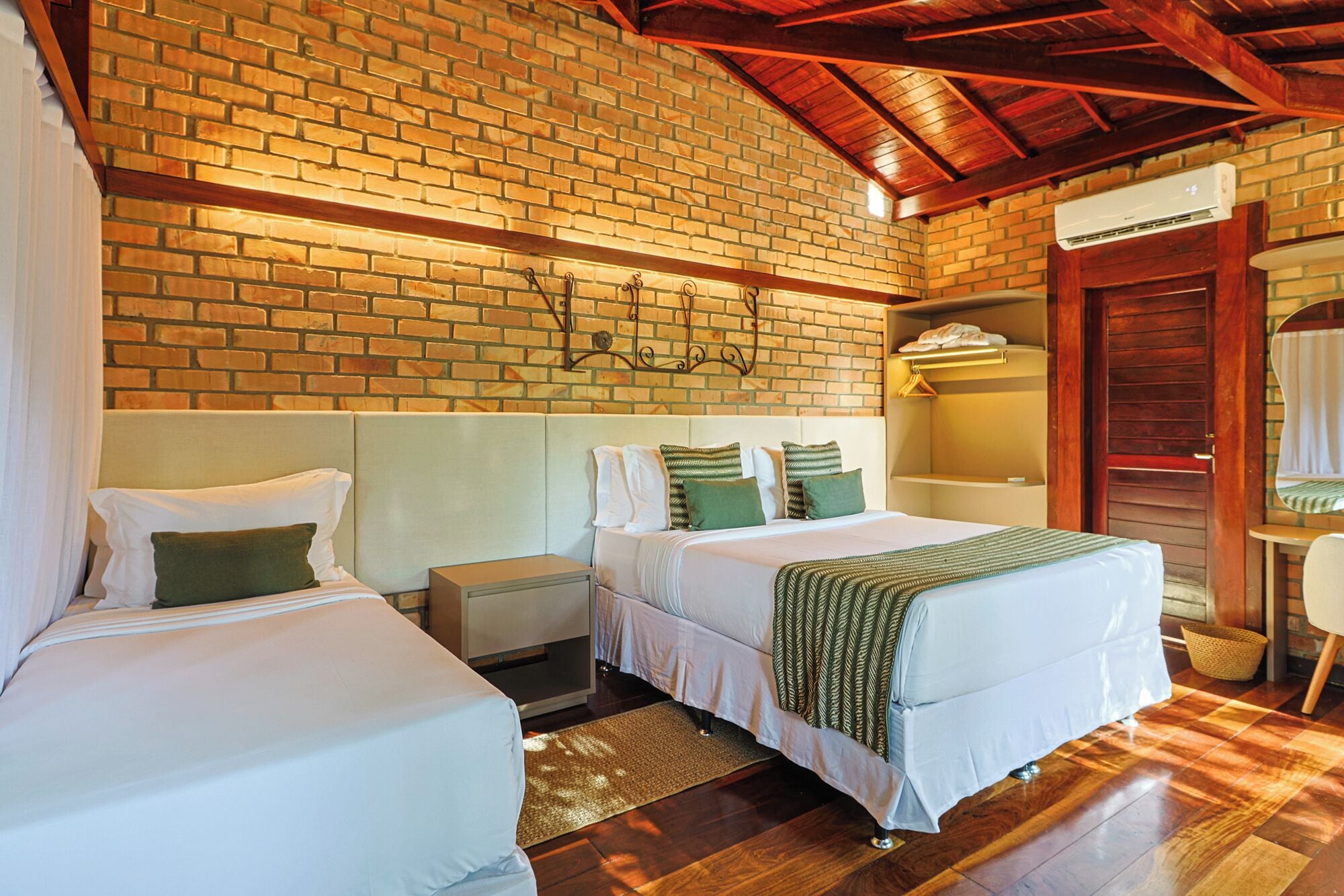 A room with two beds, a wooden ceiling, a brick wall, and a small bedside table with a lamp.