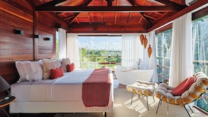Une chambre dotée d’un grand lit, d’un coin salon avec des fauteuils, d’une baignoire et offrant une vue sur la verdure grâce à de grandes fenêtres.