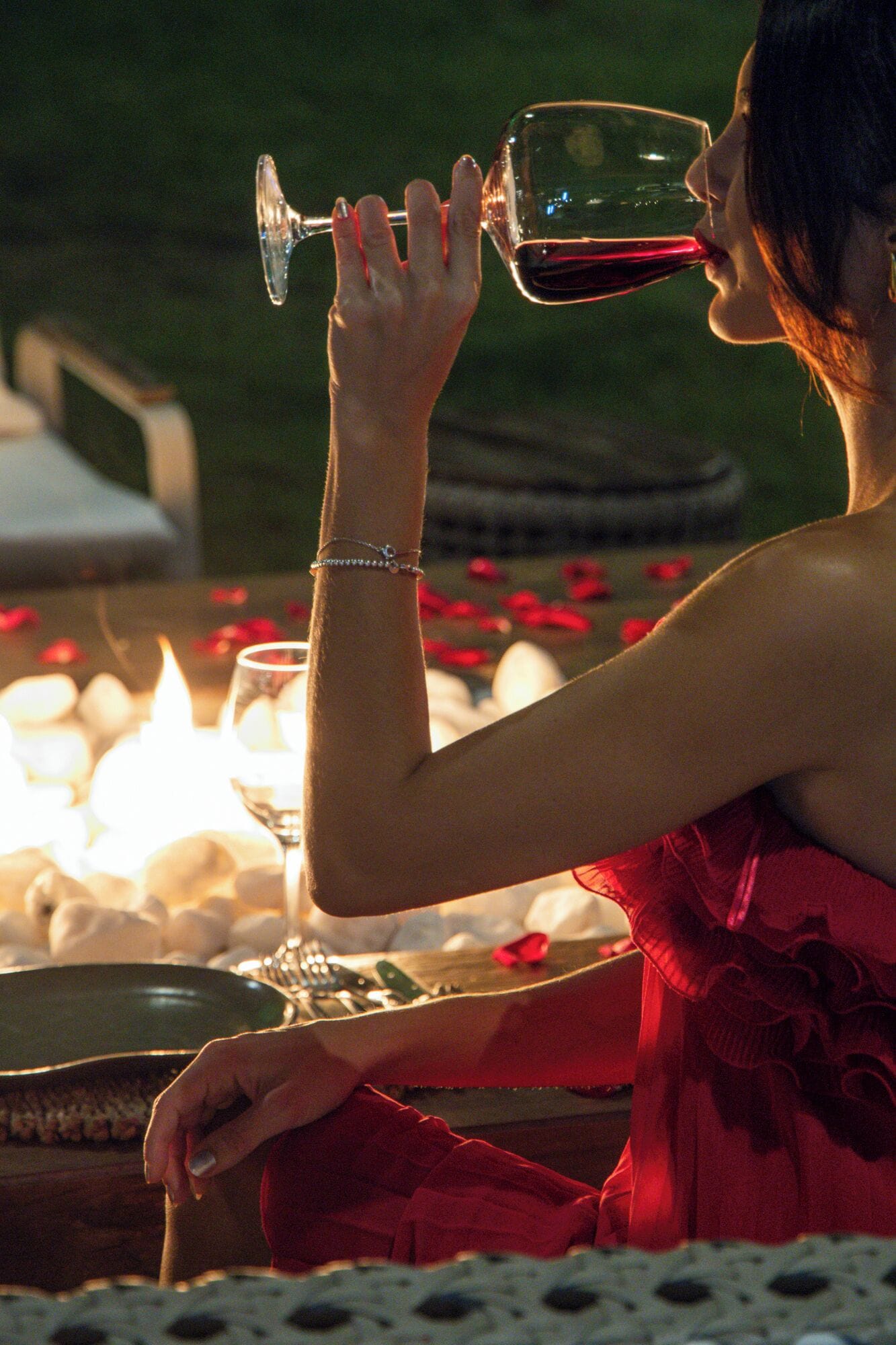 Uma pessoa de vestido vermelho sentada à mesa com velas, segurando uma taça de vinho.