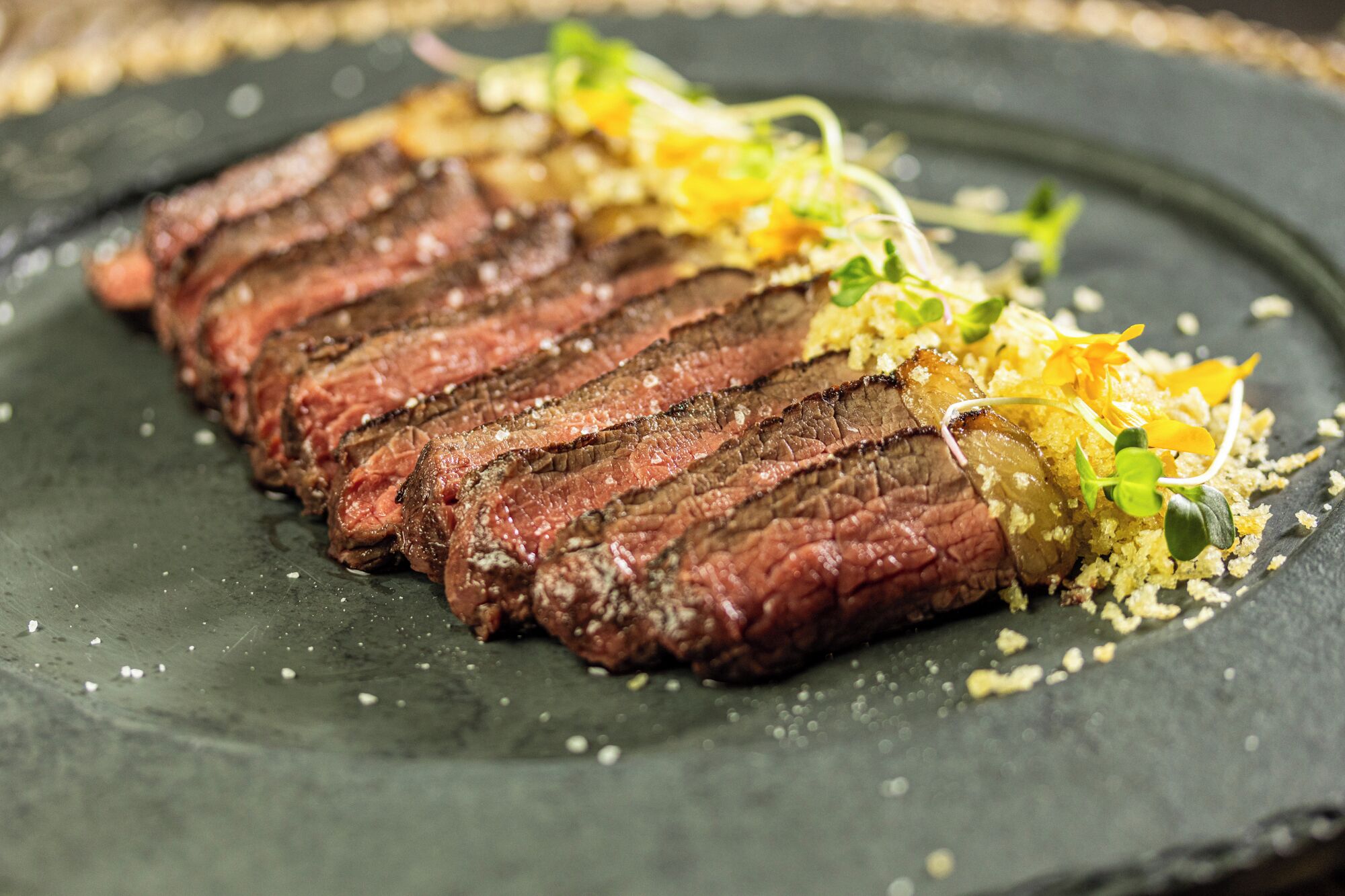 A plate of sliced steak with a garnish of greens and a yellow element, possibly a vegetable or cheese, on a dark slate plate.
