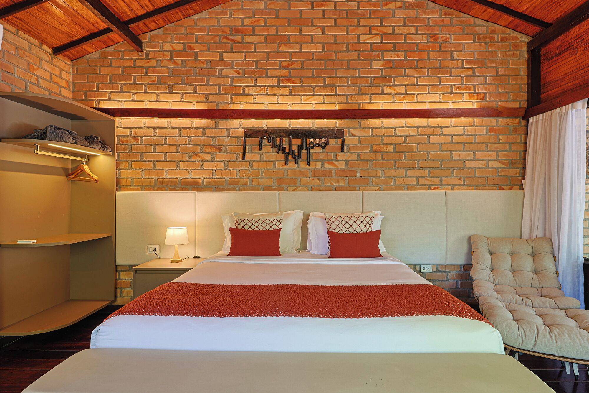 A hotel room with a brick wall, a bed with red and white bedding, a nightstand with a lamp, and a chair.