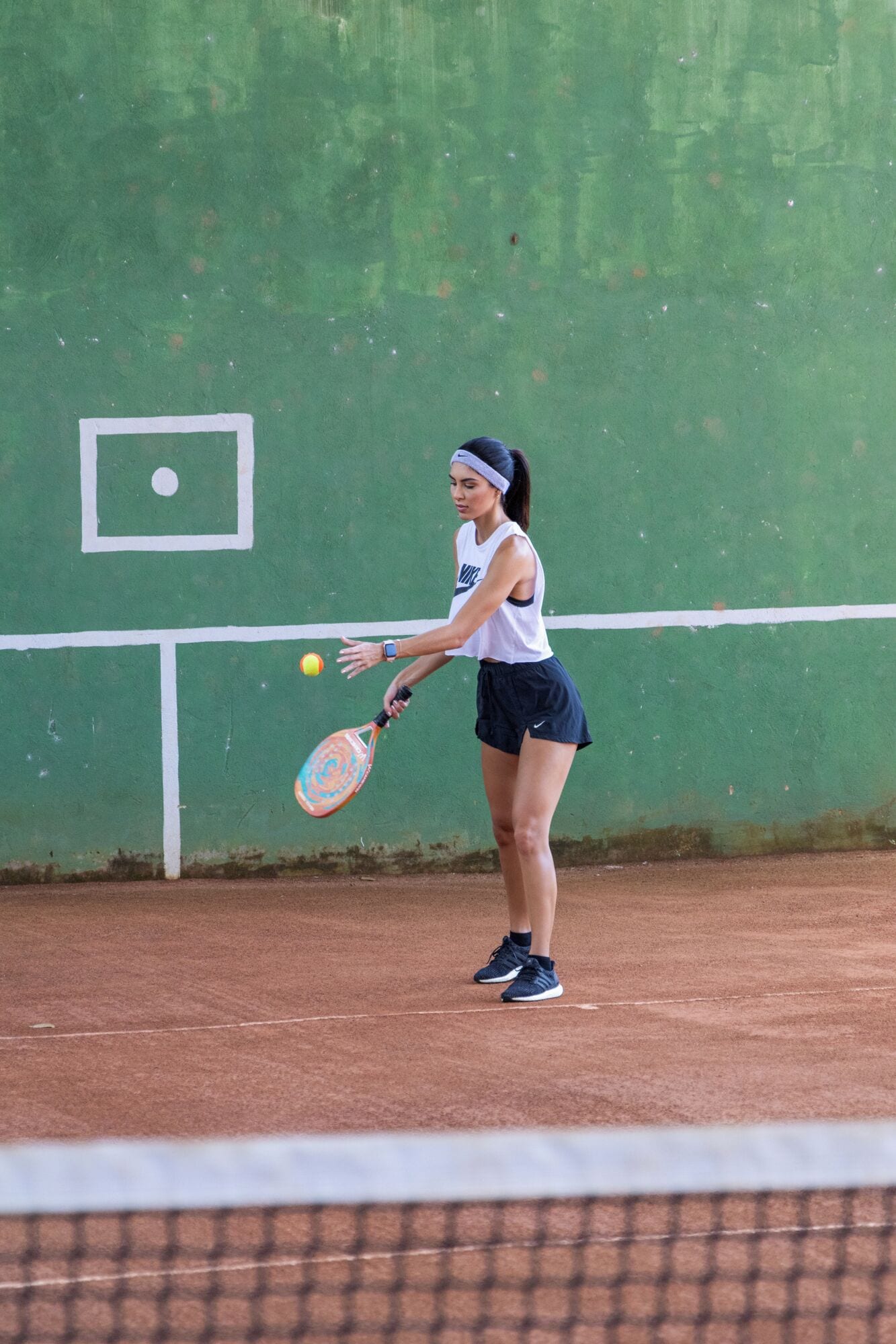 A tennis court with a player in a white top and dark shorts, holding a racket and preparing to hit a ball.