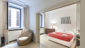 Une chambre à coucher avec un lit, une chaise en osier, une table de chevet avec une lampe, et une vue sur l’extérieur par une fenêtre.