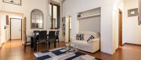 A living room with a sofa, a coffee table, a dining area with chairs, a bookshelf, and a large mirror.