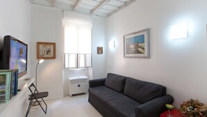 Ein Wohnzimmer mit grauem Sofa, einem Flachbildfernseher, einem kleinen Tisch mit einer Vase und einem Fenster mit Jalousien.