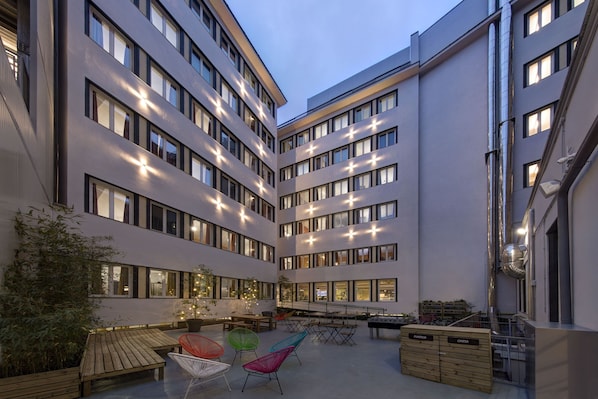 A multi-story hotel with a courtyard featuring outdoor seating, a piano, and a wooden chest of drawers.