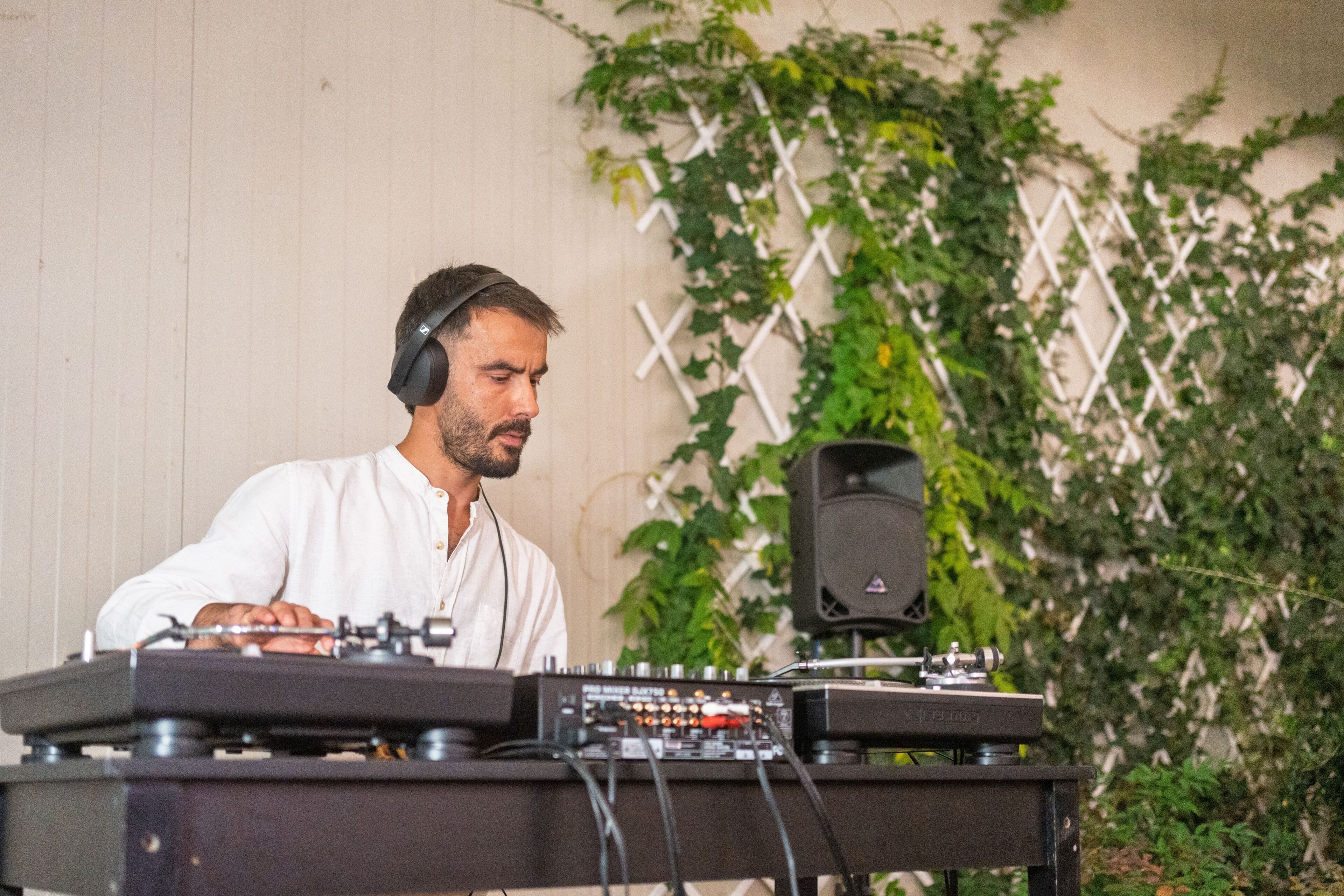A person operating DJ equipment with a speaker and a control panel, in front of a wall with a lattice design and greenery.