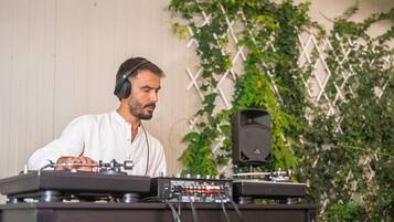 A person operating DJ equipment with a speaker and a control panel, in front of a wall with a lattice design and greenery.