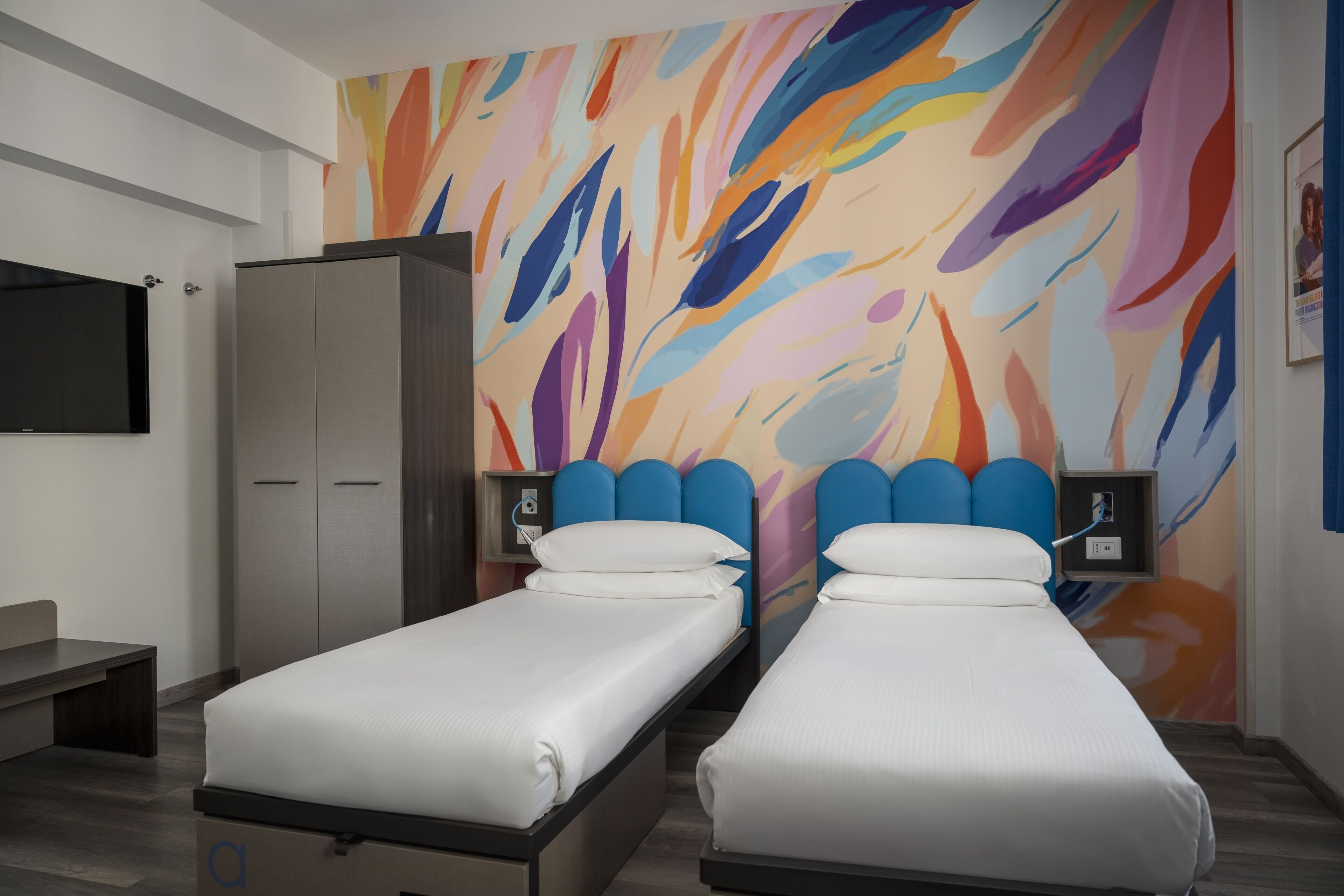 Camera d'albergo con due letti singoli, una TV, un armadio e un colorato murale astratto.