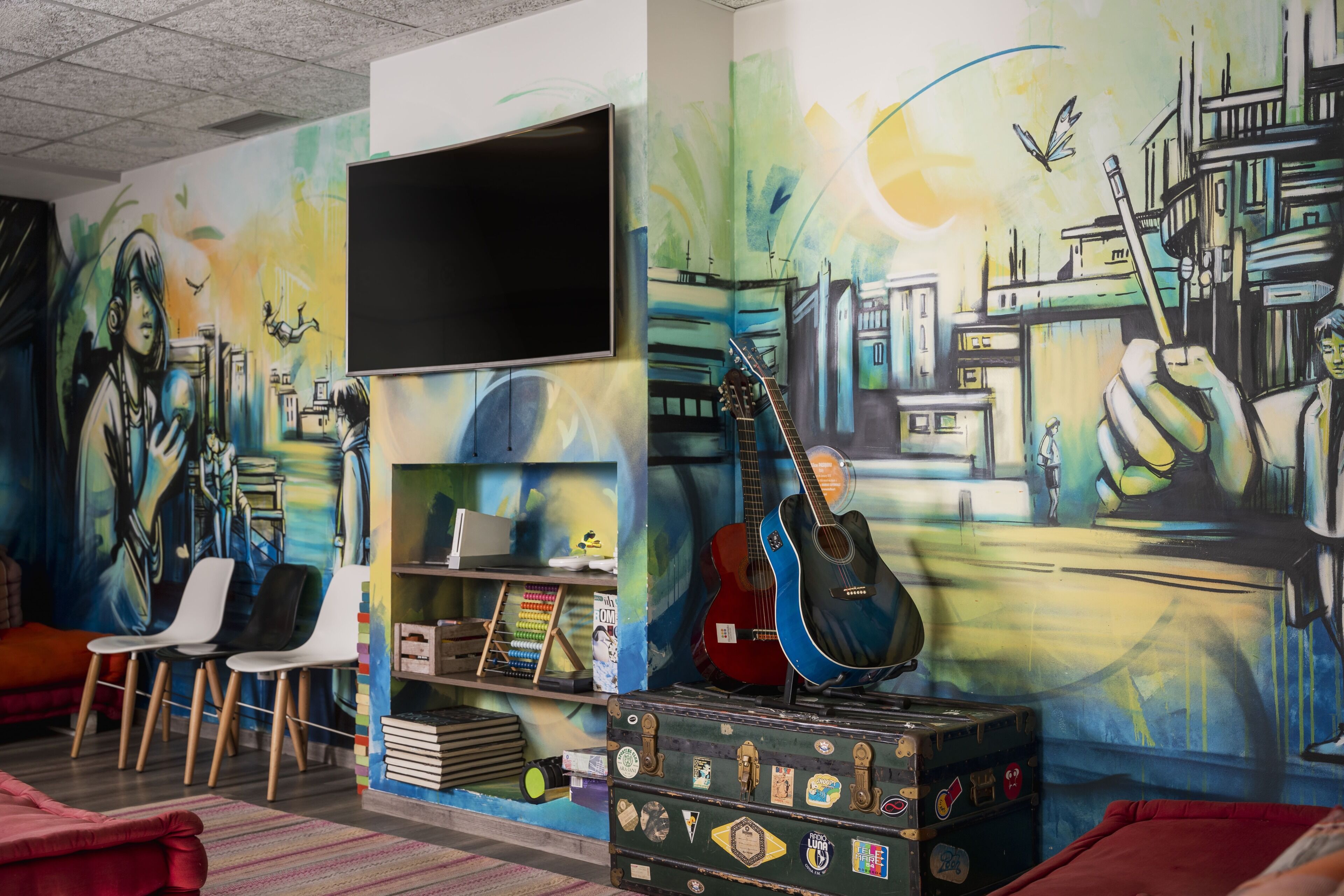 Una habitación con un mural, televisión, guitarra, sillas y una estantería.