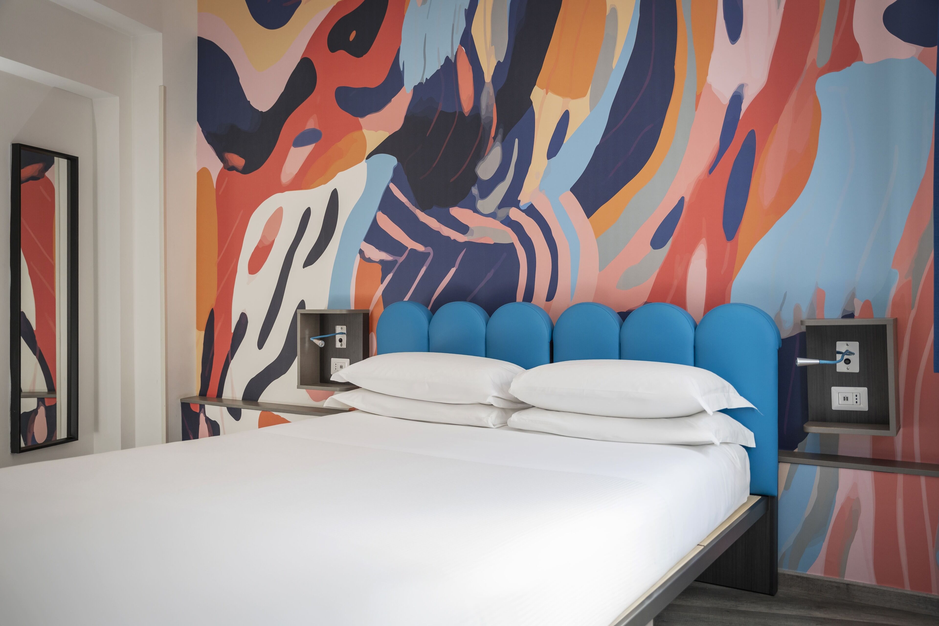 A modern hotel room with a large bed, a colorful abstract wall mural, and a nightstand with a lamp.