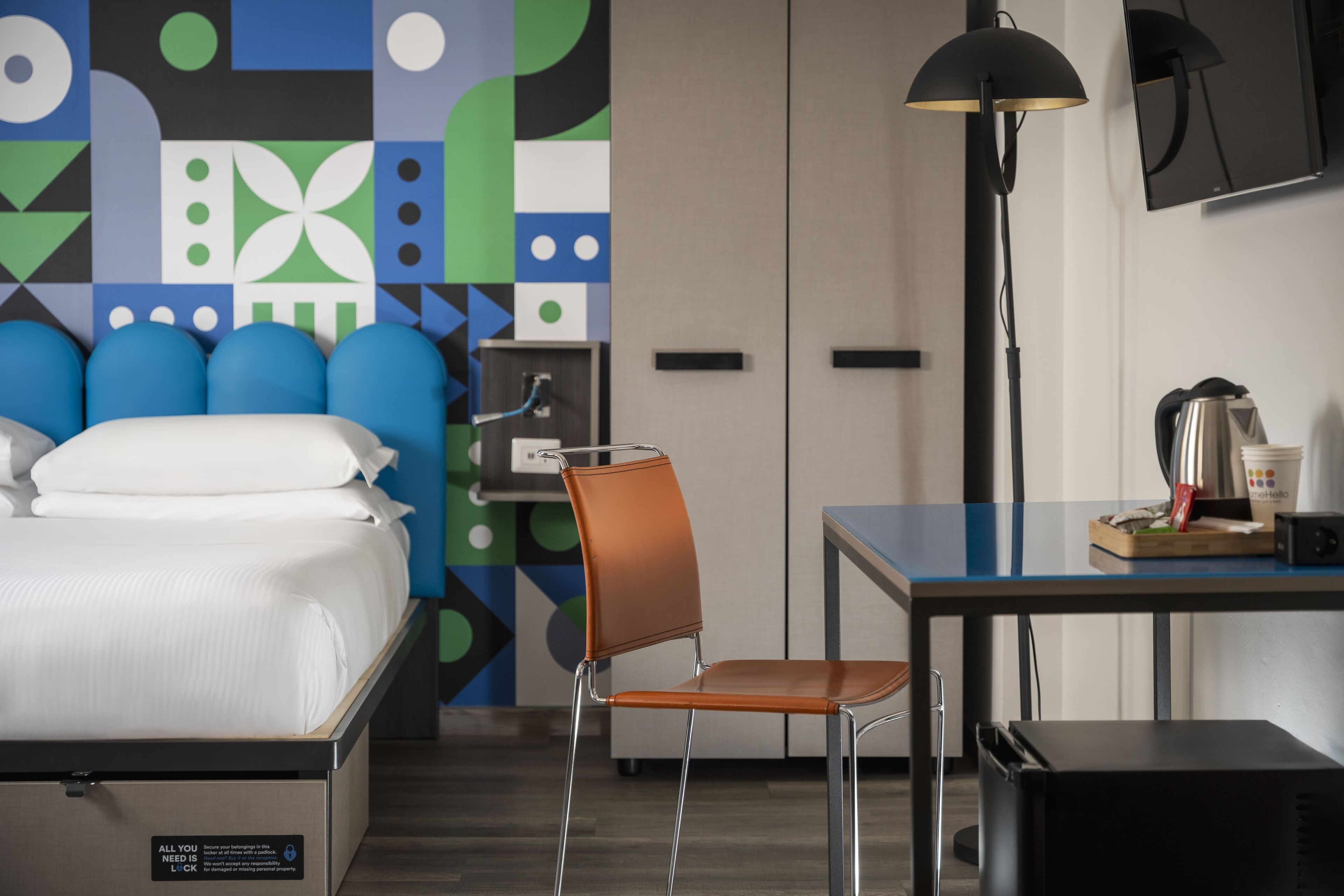 A hotel room with a bed, a desk, a chair, a lamp, a wardrobe, and a wall with a geometric pattern.