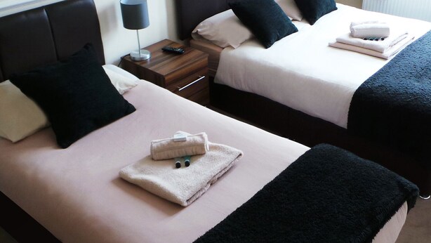 A hotel room with two beds, each with a pillow, a bedside table with a lamp, and a folded towel on the bed.