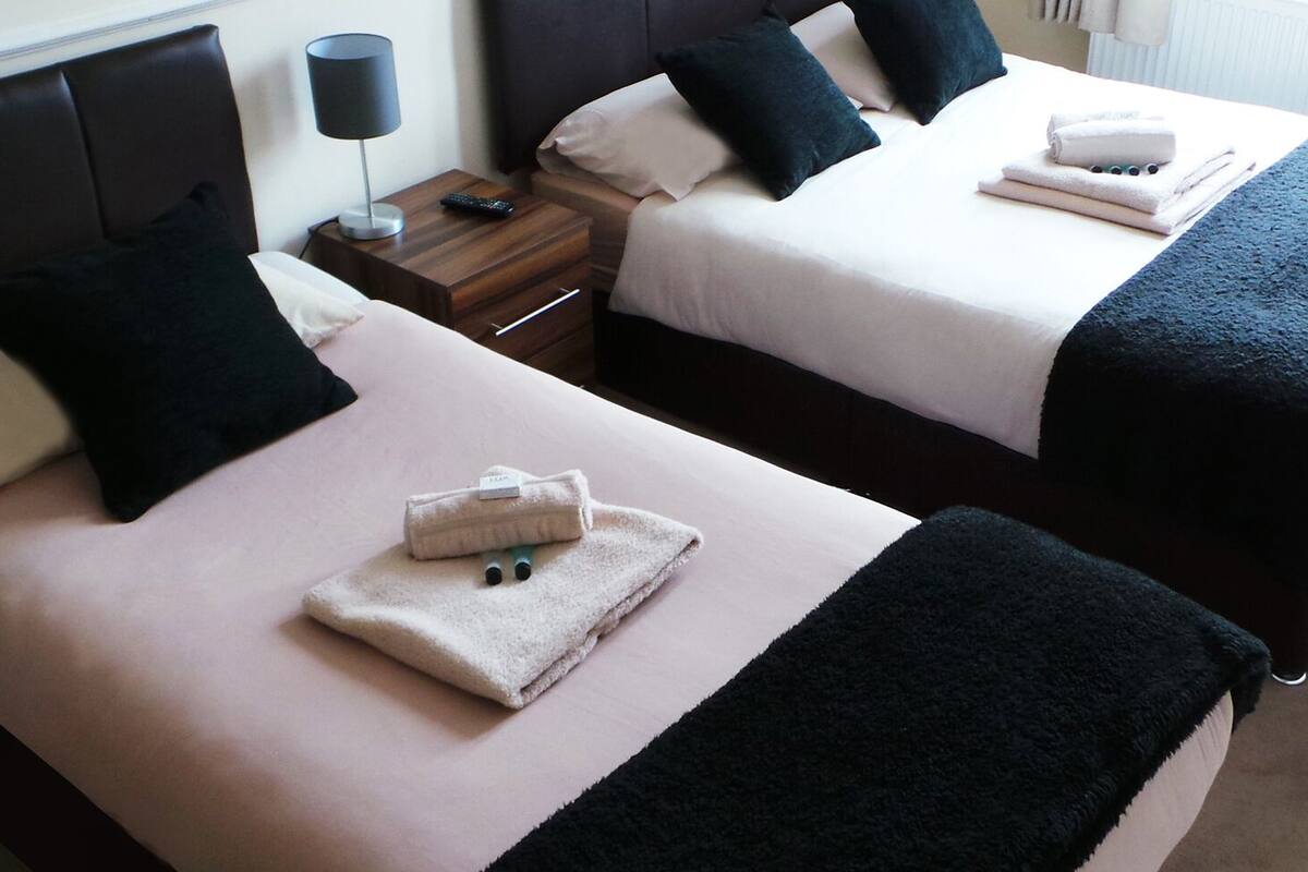 A hotel room with two beds, each with a pillow, a bedside table with a lamp, and a folded towel on the bed.