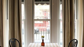 A dining area with a table set for two, a vase with flowers, and a view of the street through large windows.