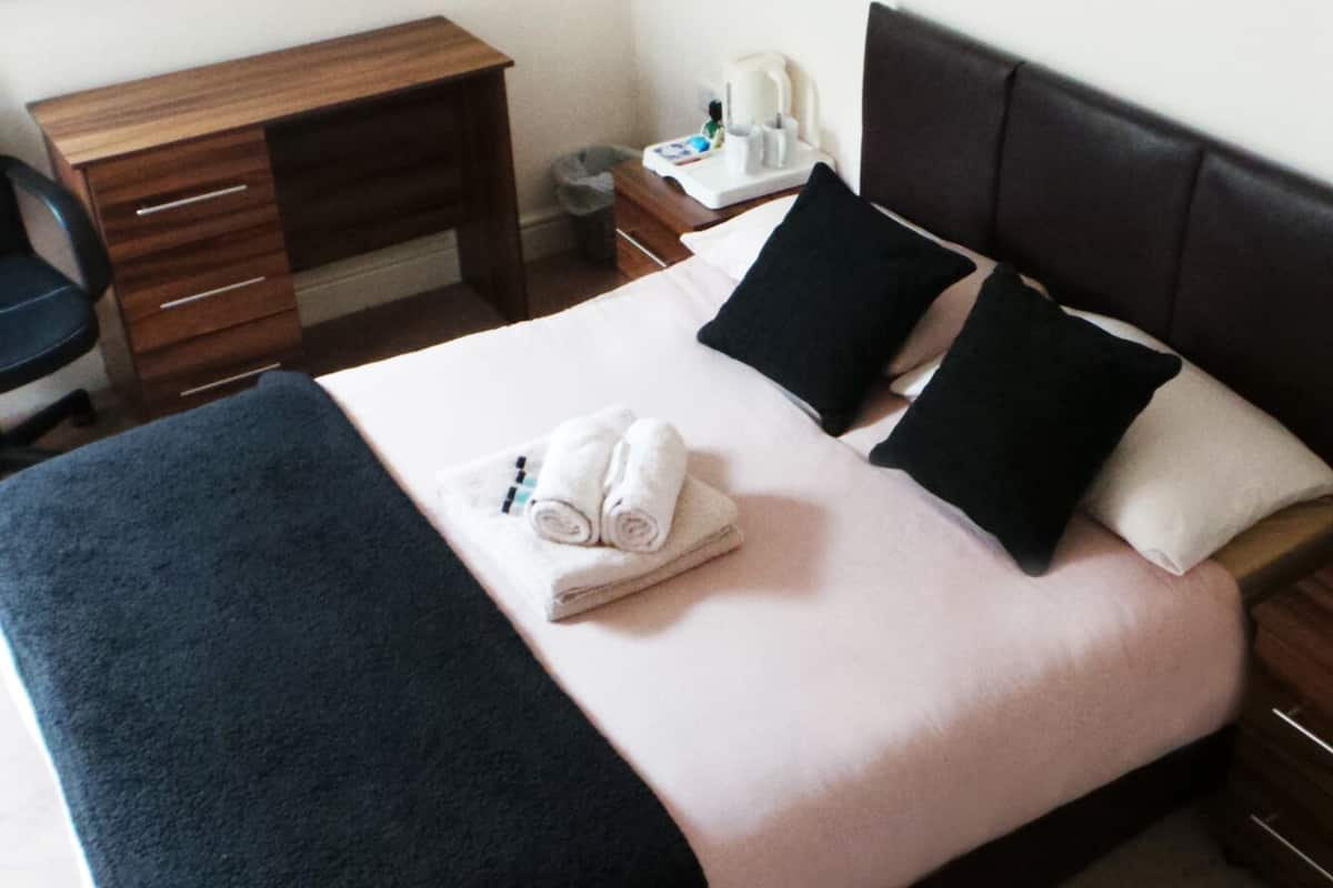 A hotel room with a bed, a desk, a chair, and a nightstand with a phone and a lamp.