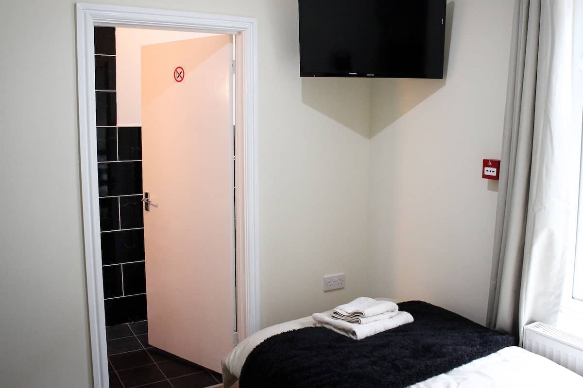 A hotel room with a bed, a TV mounted on the wall, a door leading to a bathroom, and a power outlet on the wall.