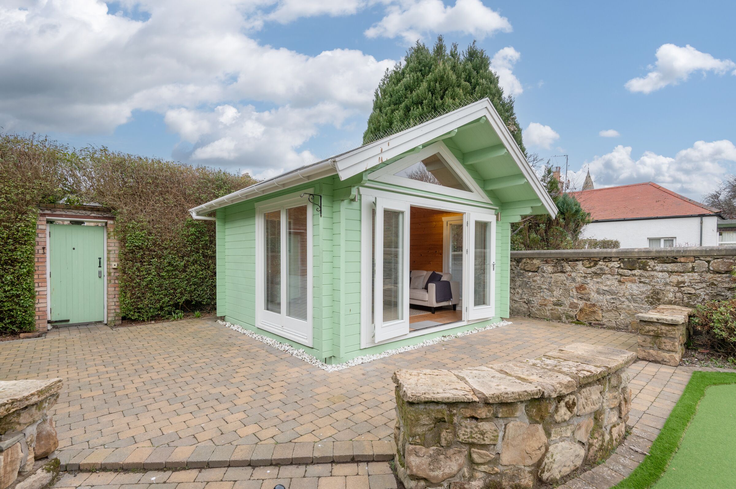 A small, light green cottage with a stone wall and a paved driveway.