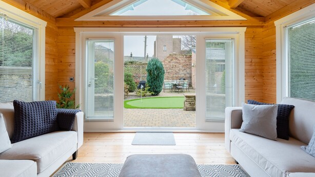 A cozy room with a wooden ceiling, two sofas, a small rug, and a view of a garden through large windows. - 138 North Street (St. Andrews)