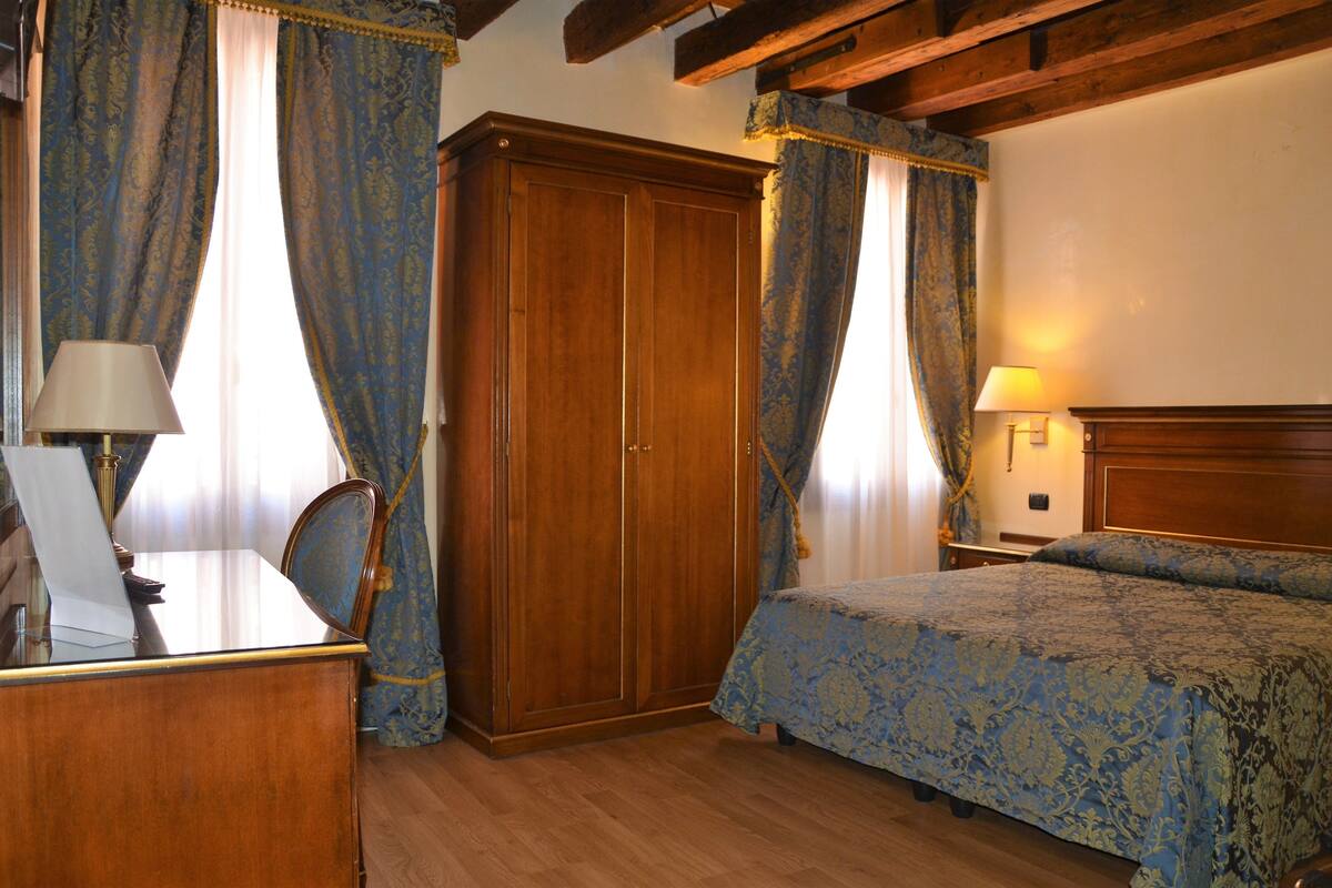 Una habitación con un armario de madera, un escritorio con lámpara, una cama con colcha estampada y cortinas.