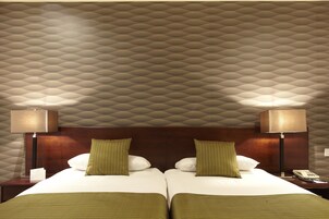 Habitación de hotel con techo estampado, dos camas con ropa de cama verde, lámparas de noche y una mesita con un teléfono.