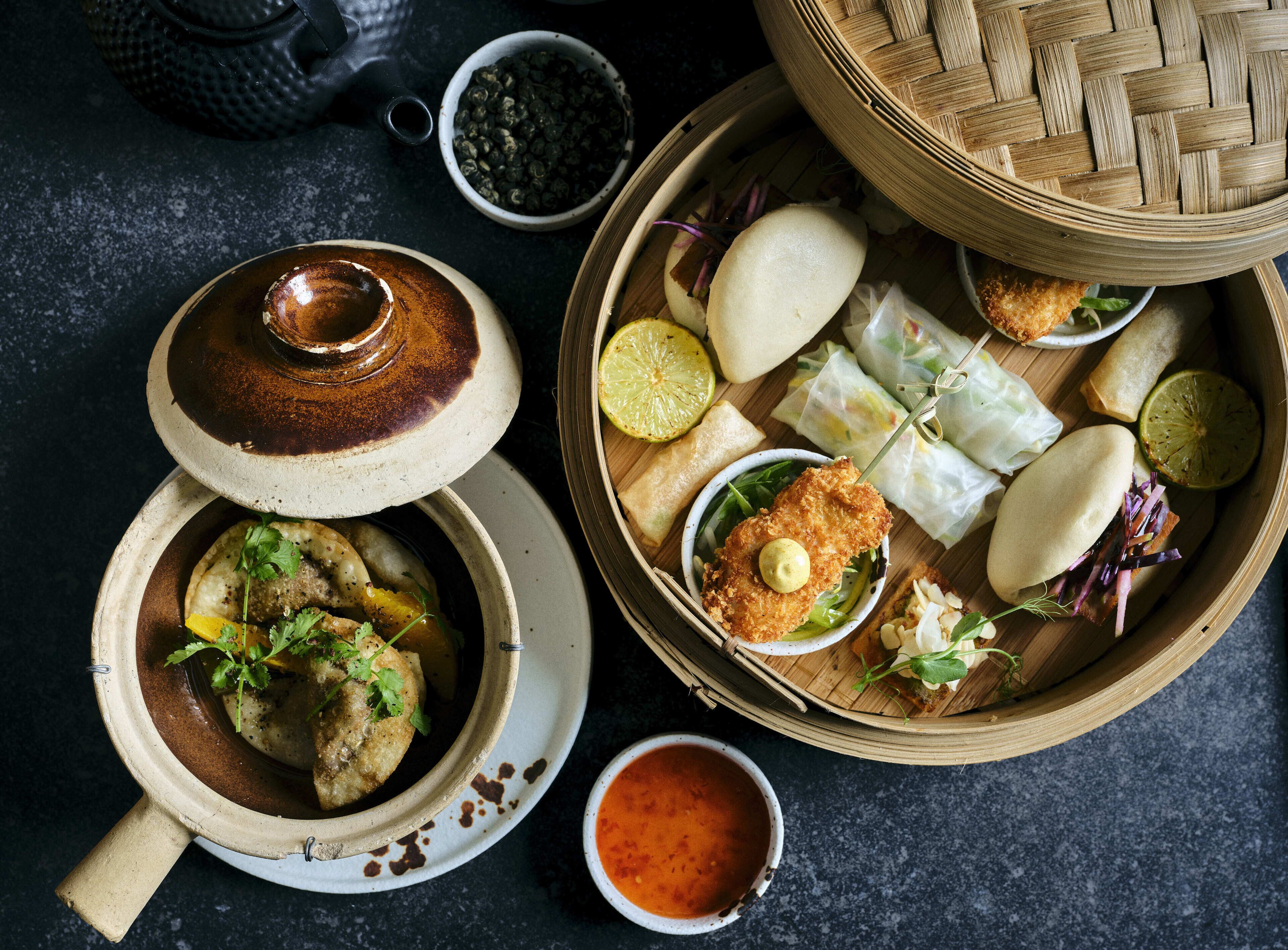 A variety of dishes including a bowl of soup, a plate with a fried item, and several spring rolls, all served on a dark surface.