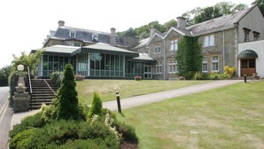 A large, historic building with a modern glass extension, surrounded by well-maintained gardens and a paved pathway. - The Manor House At Celtic Manor (Newport)