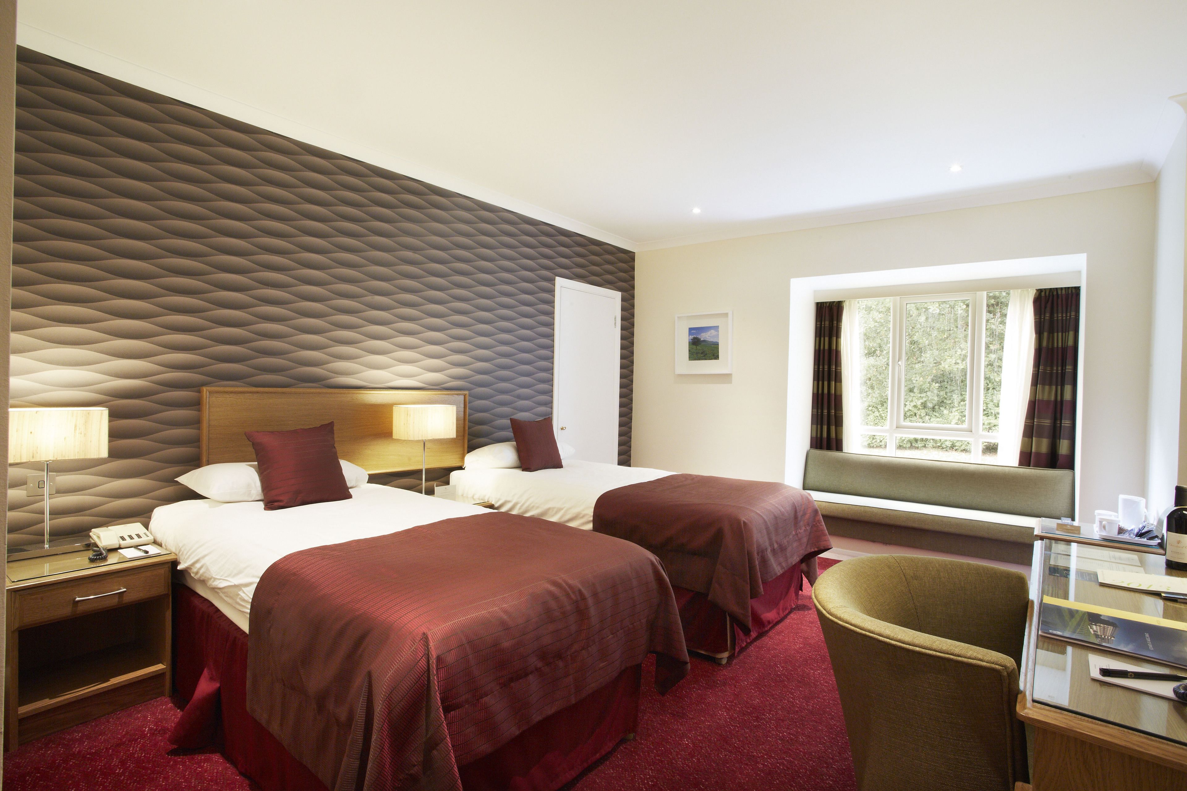 A hotel room with two beds, a desk, a chair, a window with curtains, and a textured wall.