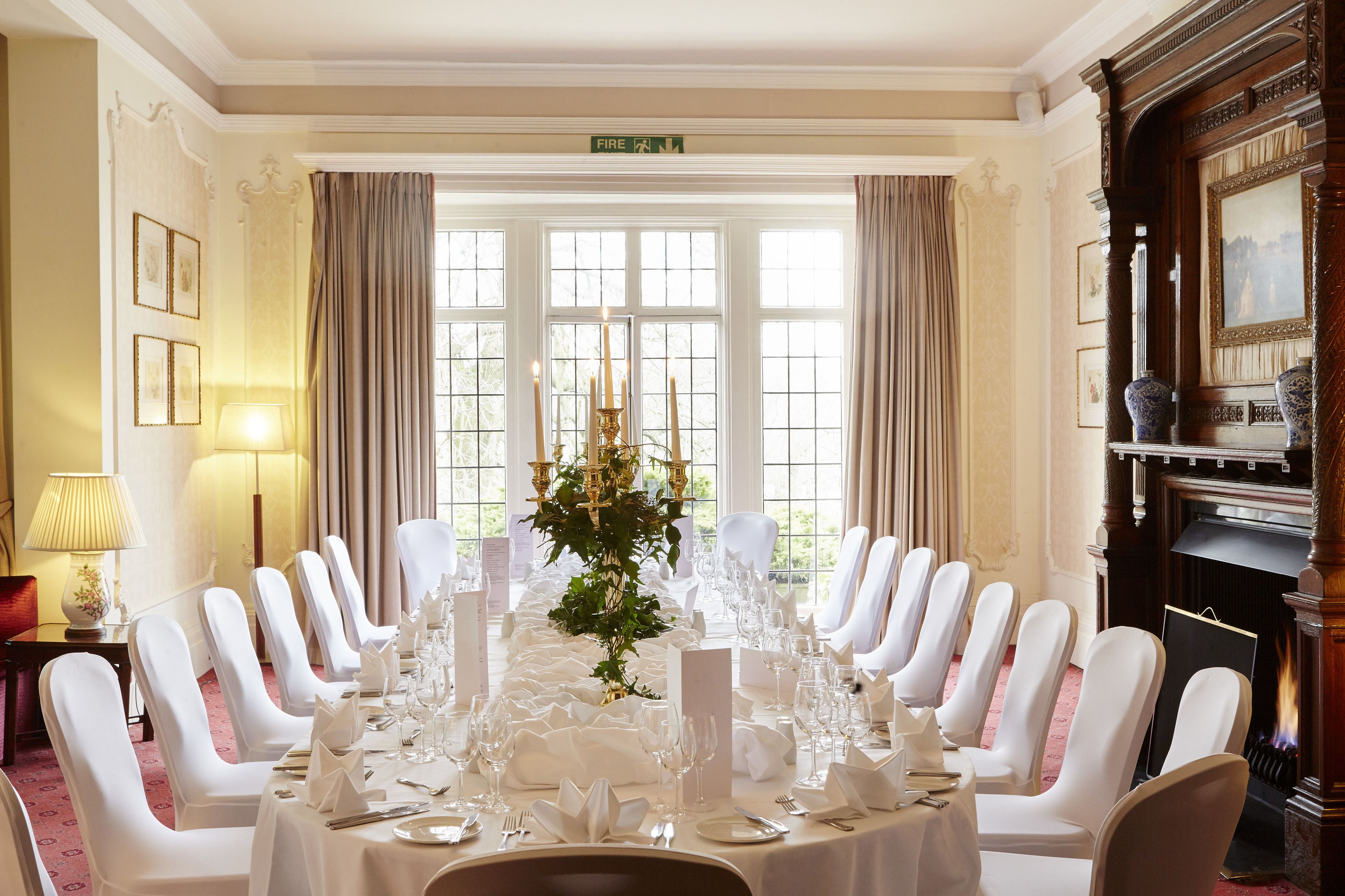 A formal dining room with a long table set for a meal, flanked by white chairs, a chandelier, and a fireplace.