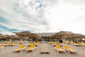 A beach with rows of yellow lounge chairs and thatched umbrellas, a wooden walkway, and a building in the background. - Boutique Citadel (Eforie)