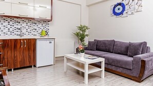 A modern living room with a grey sofa, a white coffee table, and a kitchen area with wooden cabinets and a white fridge. - MySun Hotel (Izmir)