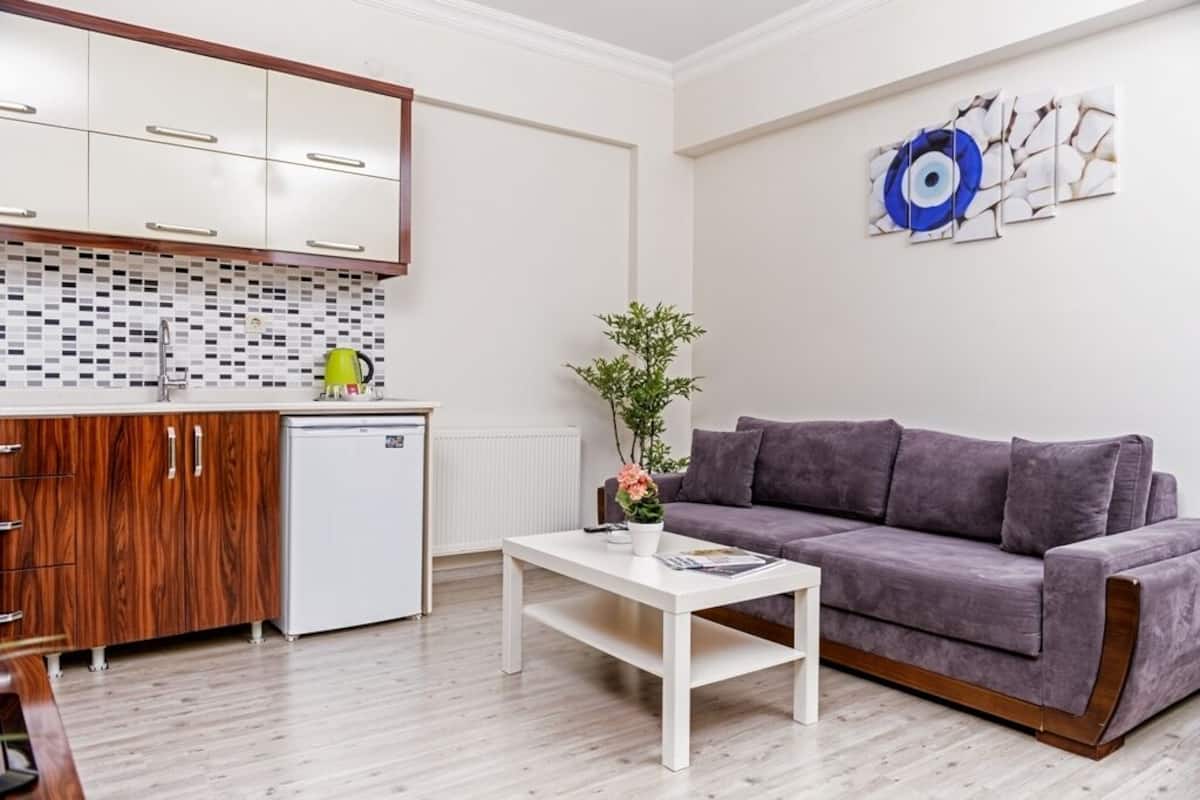 A modern living room with a grey sofa, a white coffee table, and a kitchen area with wooden cabinets and a white fridge.
