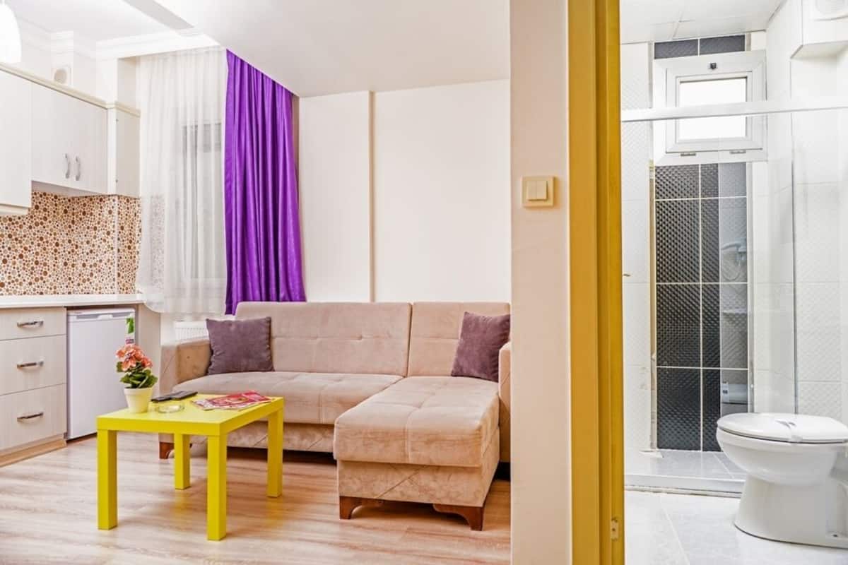 A modern living room with a sofa, a yellow table, and a kitchen area with a white cabinet and a small fridge.