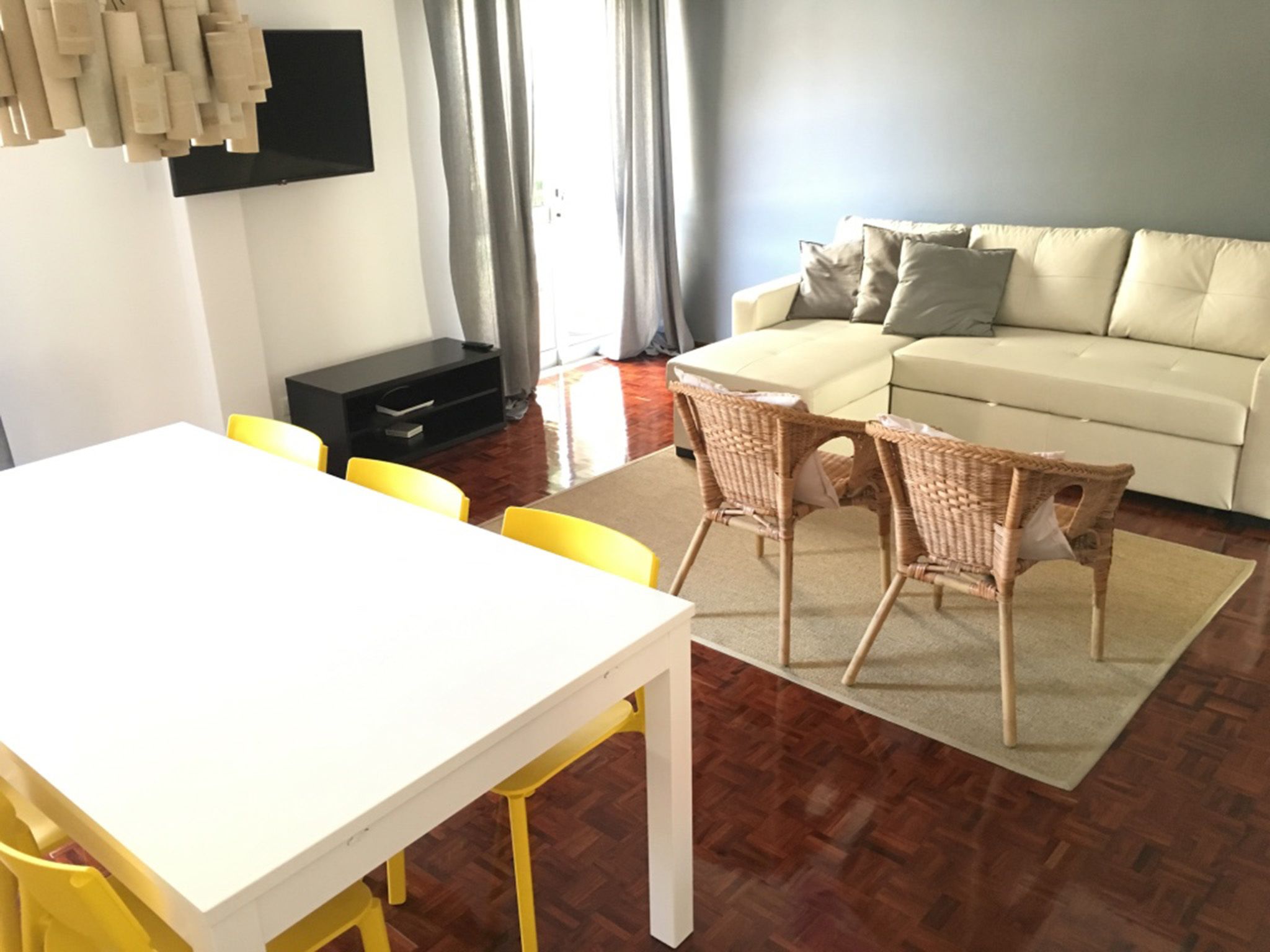 Een moderne woonkamer met een witte bank, gele stoelen, een eettafel en een flatscreen televisie aan de muur.