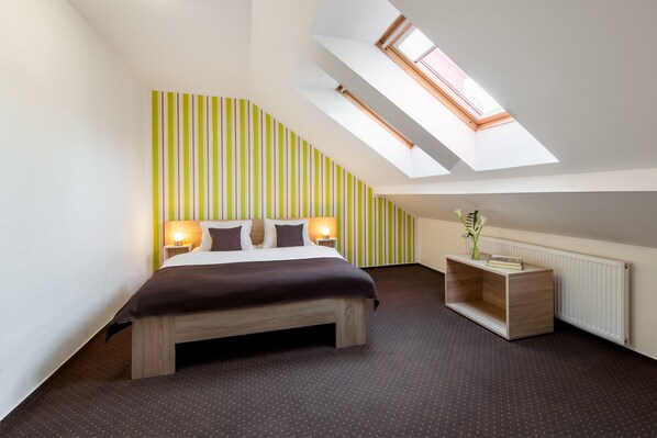 A modern bedroom with a sloped ceiling, a bed with a wooden frame, a nightstand with a lamp, a small wooden table with a vase, and a radiator. - Hotel Bon (Tanvald)