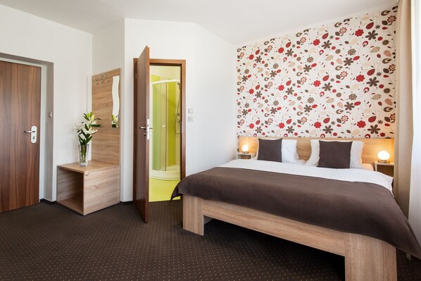 A hotel room with a wooden bed, a nightstand with a lamp, a mirror, a wooden bench, and a door leading to a bathroom. - Hotel Bon (Tanvald)
