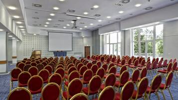Un salón de conferencias con filas de sillas rojas, una pantalla de proyector y ventanas amplias.