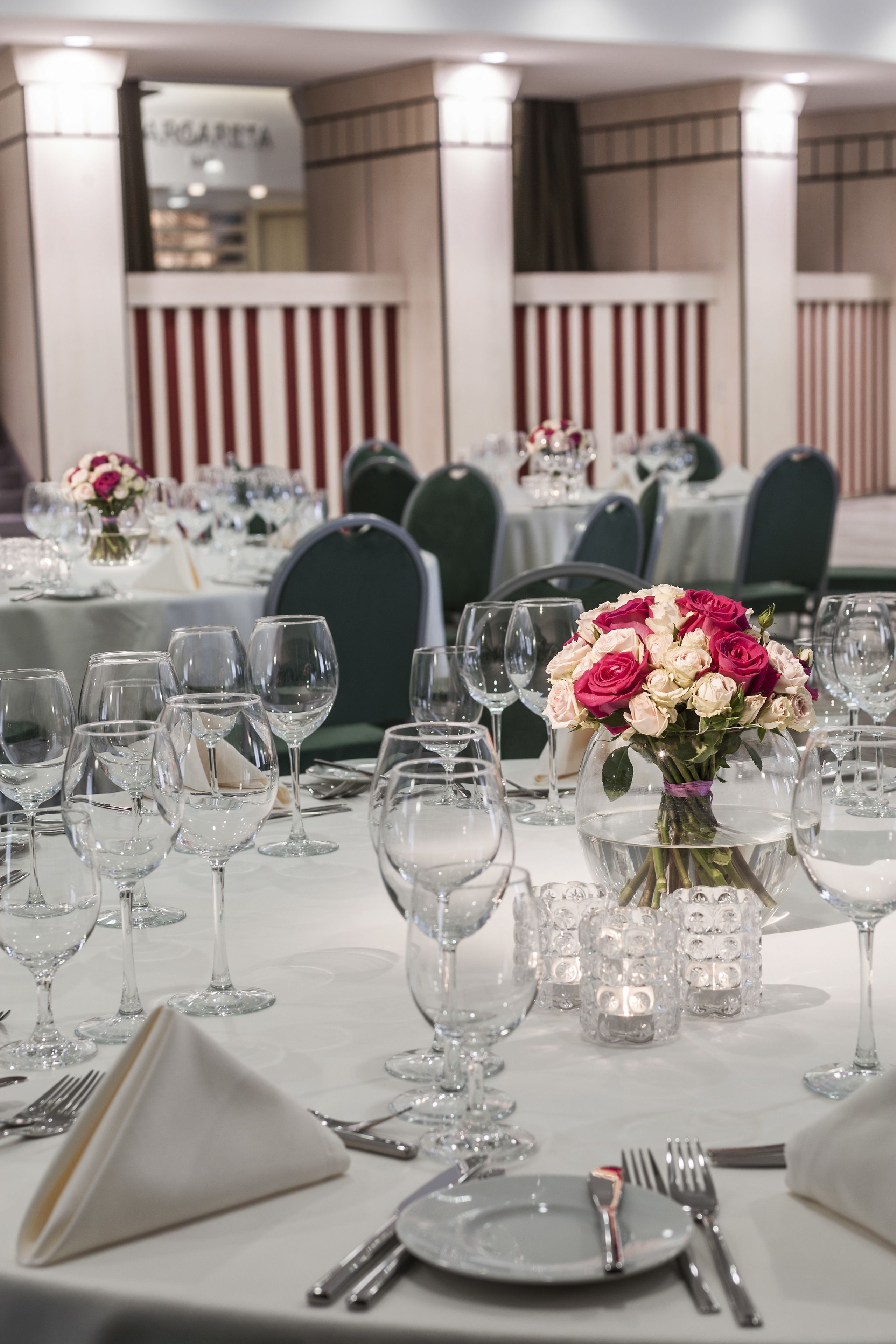A banquet hall with round tables set for a formal event, featuring clear glassware and a centerpiece of pink and white flowers.