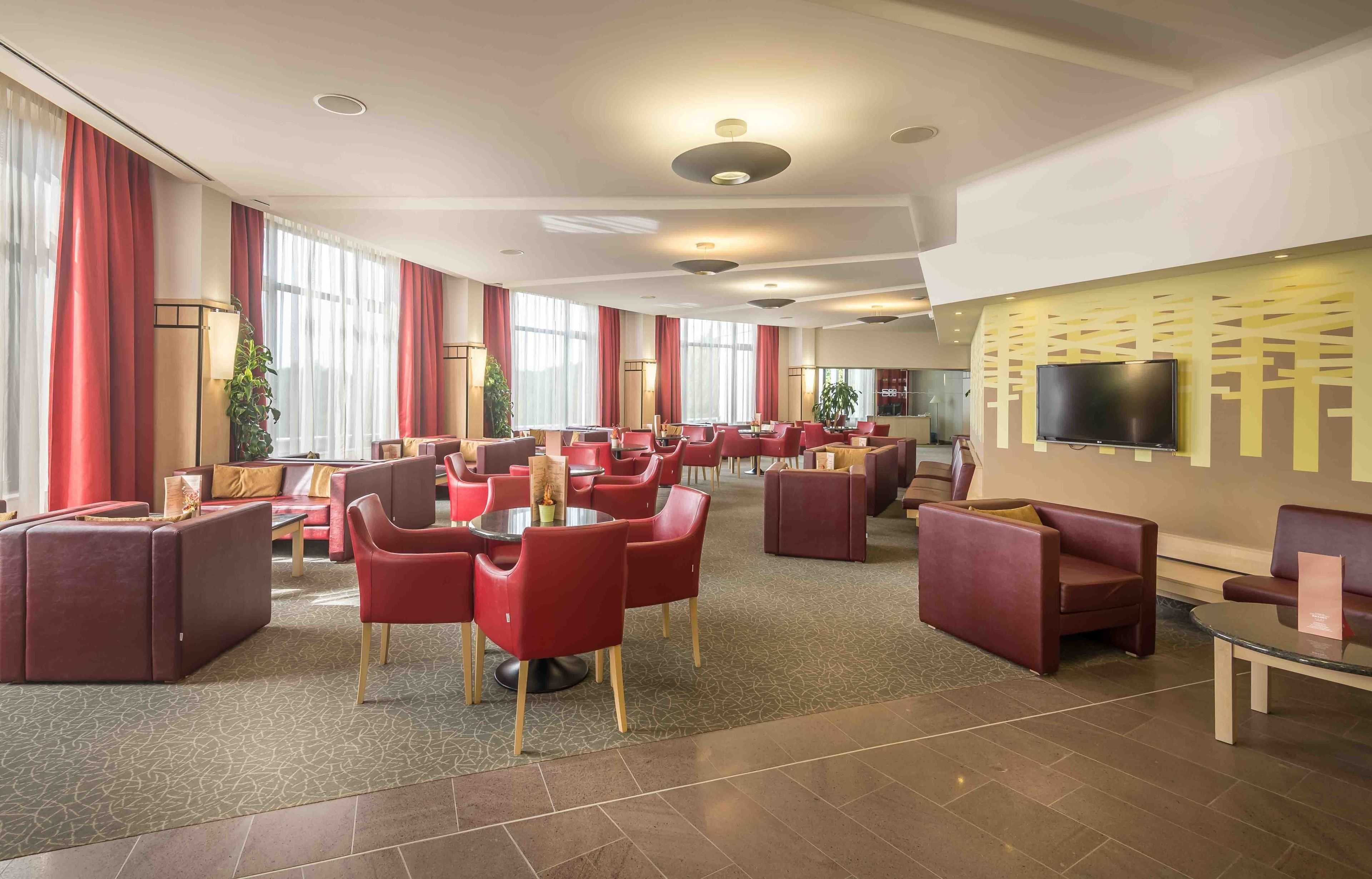 A spacious hotel lobby with red seating, a flat-screen TV mounted on the wall, and large windows with red curtains.
